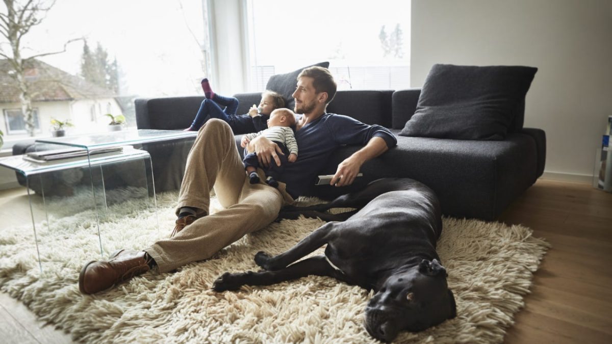 Man sits in the living room on the floor in front of the television with his kids and the family dog.